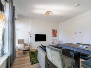 Un salon moderne doté d’une grande fenêtre, d’un téléviseur à écran plat, d’une table à manger et de deux fauteuils.