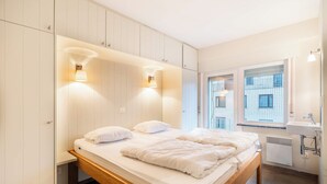 A modern bedroom with a wooden bed, white walls, and built-in cabinets. There are two wall-mounted lights and a window with a view of buildings. - Apartment by Belgian Coast With Beach Views (Koksijde)