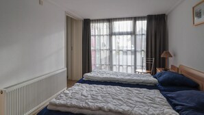 Una camera d'albergo con due letti, un termosifone, una finestra con tende, una sedia e una lampada.