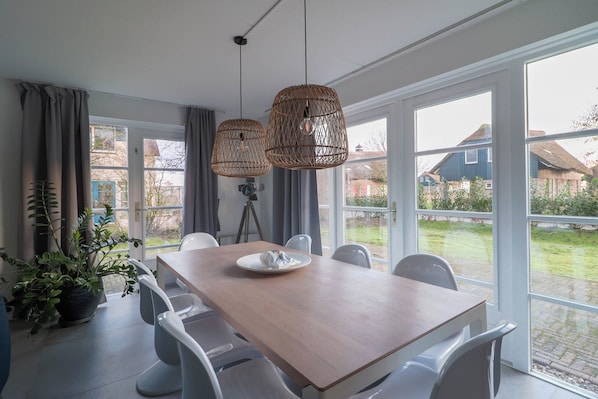 Un comedor con mesa de madera, sillas blancas y grandes ventanales con vistas a casas.