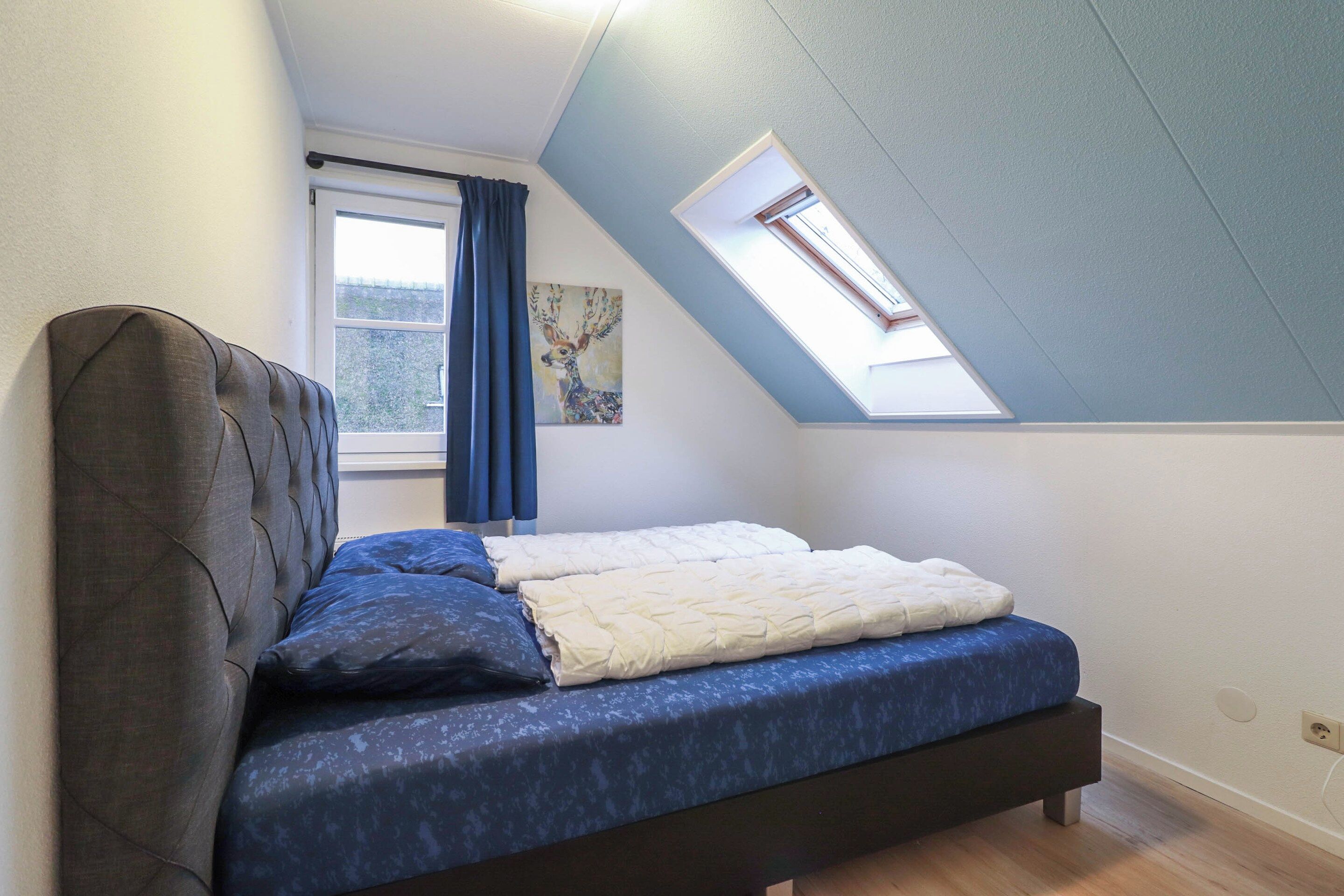 Une chambre avec un plafond en pente, un puits de lumière, un lit avec une literie bleue et blanche, et une fenêtre avec des rideaux bleus.