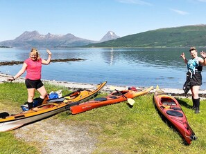 Zwei Kajaks und ein Stand-Up-Paddleboard am Ufer eines Sees, im Hintergrund Berge.