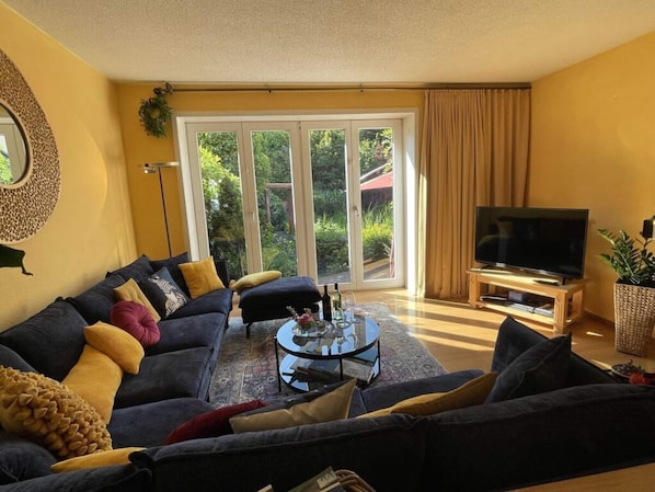 A living room with a dark-colored sofa set, a coffee table, a flat-screen TV on a wooden stand, and a sliding glass door leading to a garden.