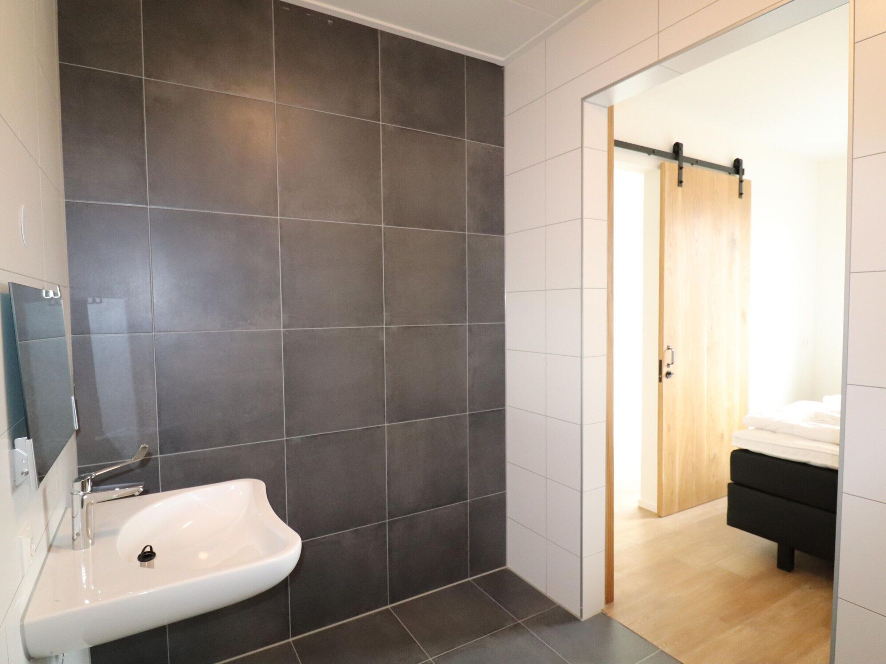 Une salle de bain moderne avec des carreaux foncés, un lavabo blanc et une porte en bois donnant sur une chambre à coucher équipée d’un lit et d’une table de chevet.