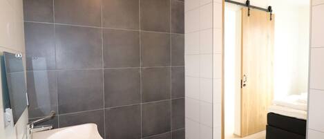 Une salle de bain moderne avec des carreaux foncés, un lavabo blanc et une porte en bois donnant sur une chambre à coucher équipée d’un lit et d’une table de chevet.