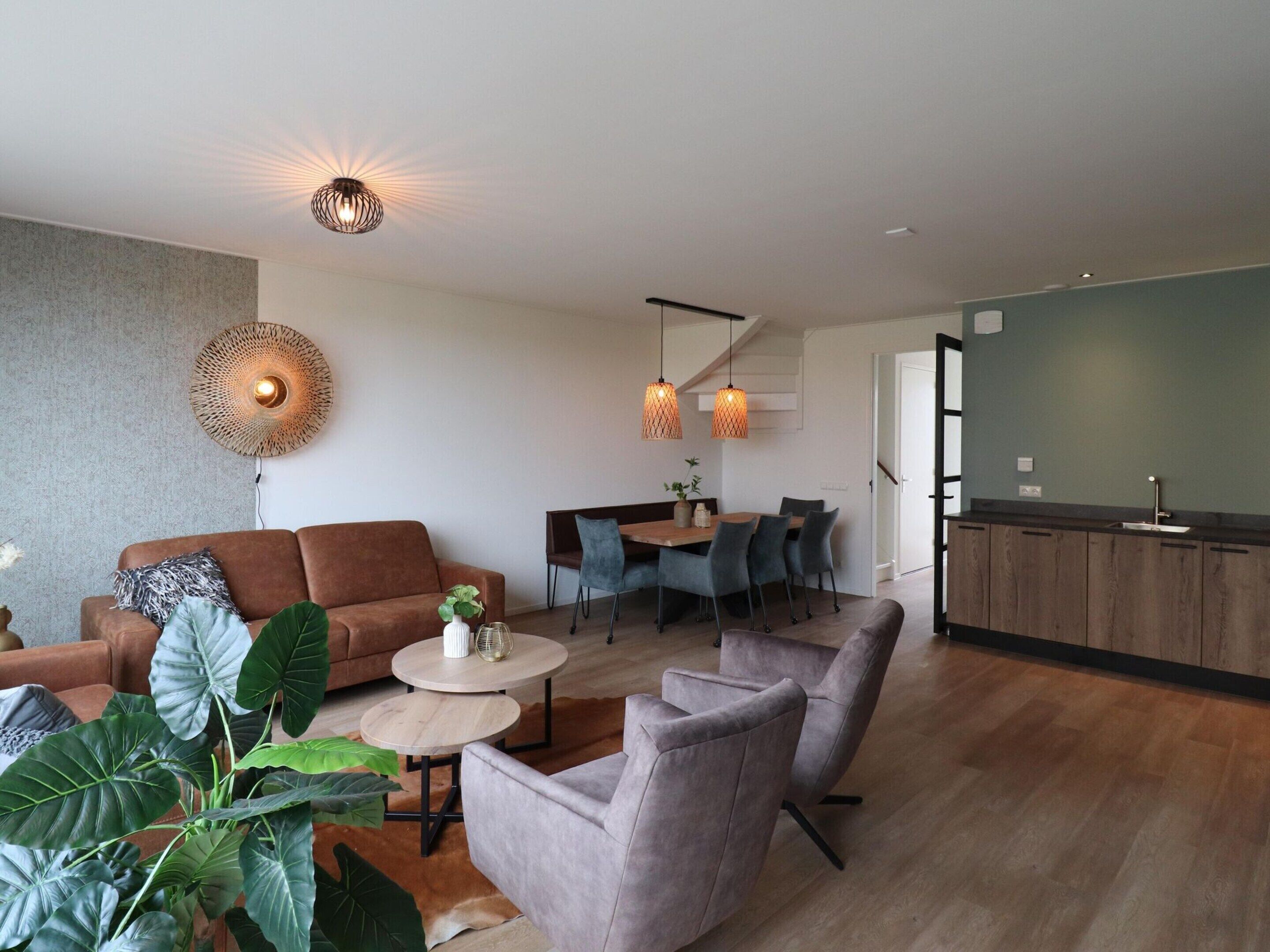 A modern living room with a sofa, armchairs, a coffee table, and a dining area with chairs. There is a kitchen area with a counter and cabinets.