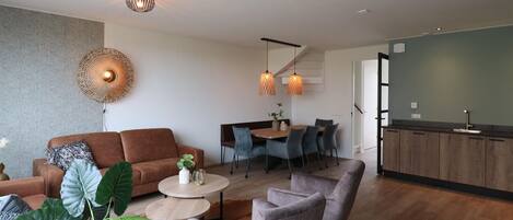 A modern living room with a sofa, armchairs, a coffee table, and a dining area with chairs. There is a kitchen area with a counter and cabinets.