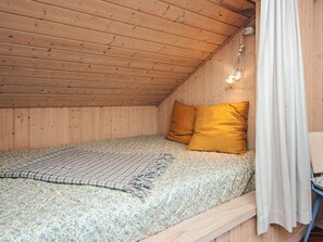 A small, cozy room with a sloped ceiling, wooden walls, a bed with a floral bedspread, two pillows, and a white curtain. - 6 Person Holiday Home in Norre Nebel (Norre Nebel)