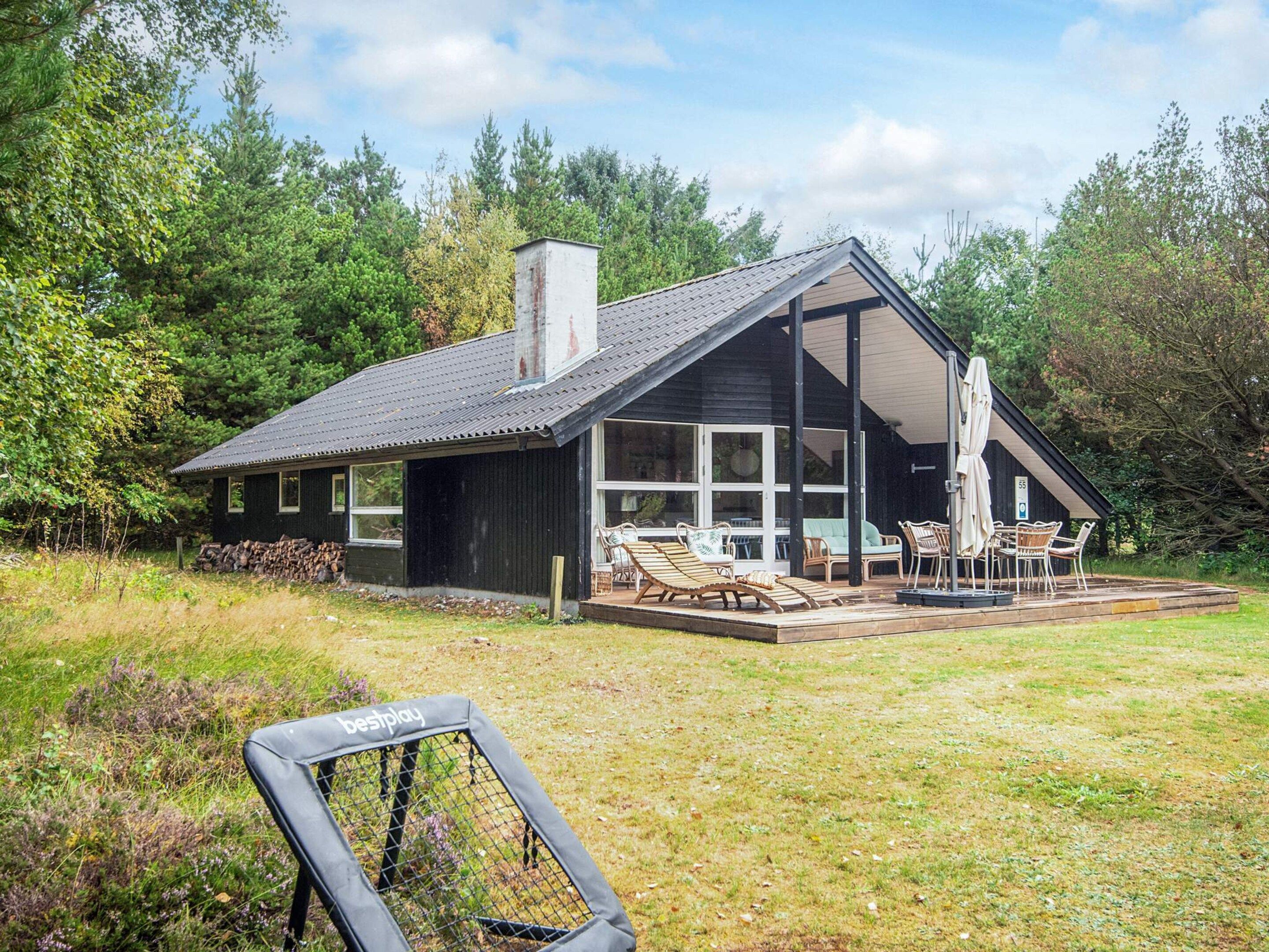A modern house with a large deck, outdoor seating, and a view of the surrounding forest.