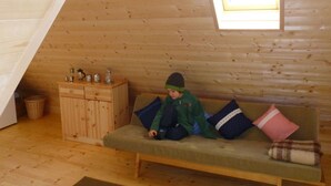Ein gemütlicher Raum mit schrägverdeckter Holzbalkendecke, einem kleinen Fenster, einer Couch mit Kissen und einem Holzkasten mit Gegenständen darauf.
