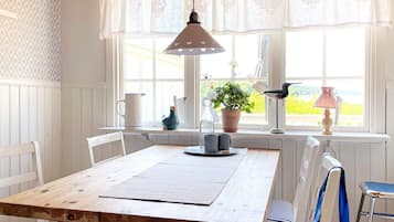 Un comedor con mesa de madera, sillas blancas, una lámpara colgante y vistas a través de una ventana con cortinas de encaje.