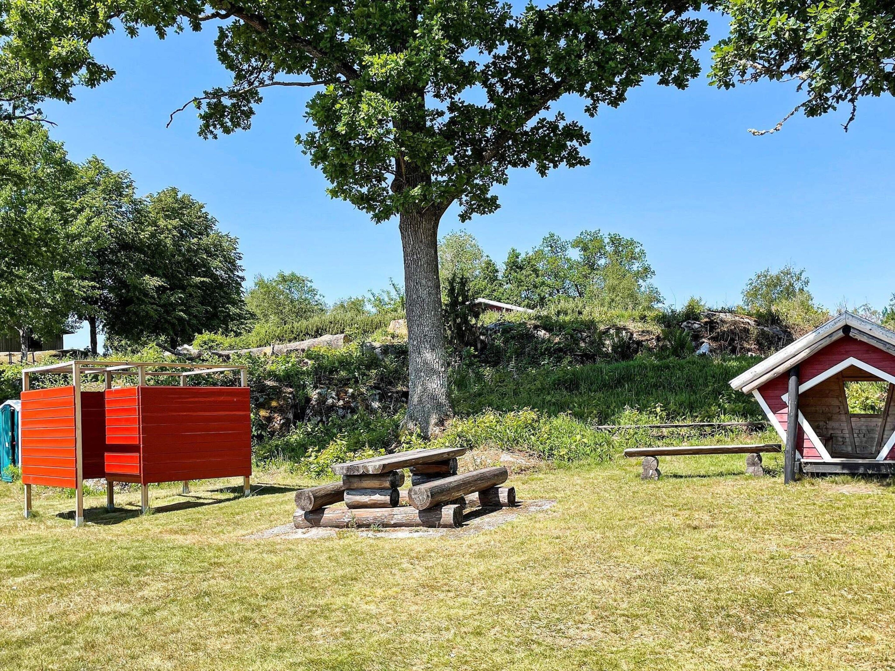 A picnic area with a wooden bench and a small red structure, surrounded by trees and a grassy field.