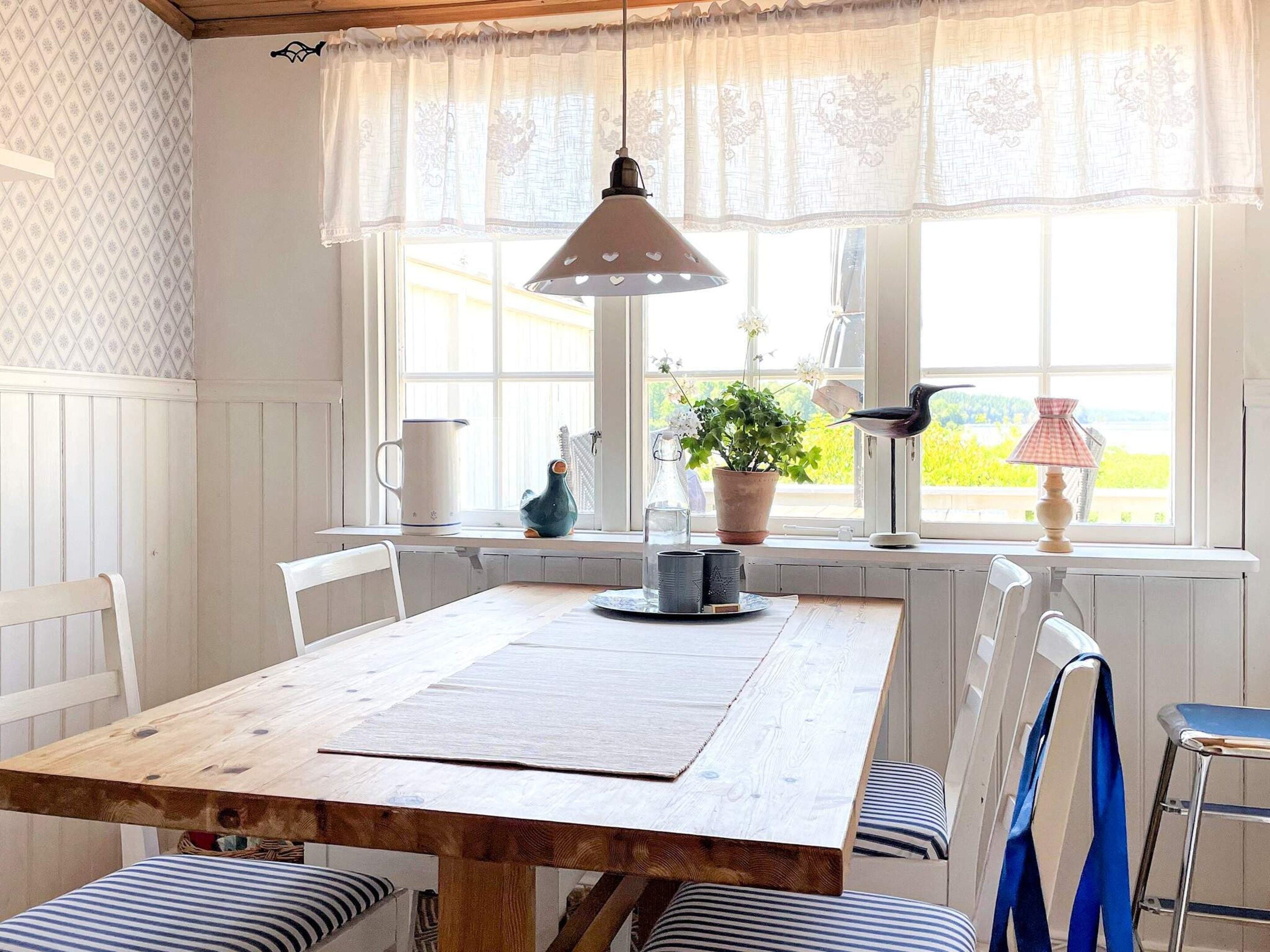 A dining area with a wooden table, white chairs, a hanging pendant light, and a view through a window with a lace curtain.