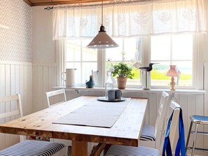 A dining area with a wooden table, white chairs, a hanging pendant light, and a view through a window with a lace curtain. - 4 Star Holiday Home in Aplared (Aplared)