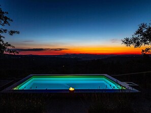 A rectangular pool with a single candle in the center, surrounded by grass and trees, with a sunset view in the background.