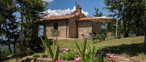 A stone building with a tiled roof, a small bell tower, and a covered patio area with two lounge chairs.