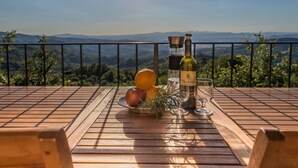 Ett träbord med en vinflaska, ett glas och en skål med frukt på en balkong med utsikt över bergen.