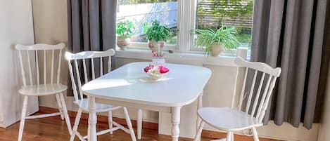 A dining area with a white table and four white chairs, a vase with flowers, and a view of greenery outside.