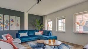A modern living room with a blue sectional sofa, a round coffee table, and a large window with a view of a building. - Villa With hot tub and Sauna, on a Holiday Park (Ossenisse)