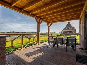 A wooden deck with a table and chairs, overlooking a river and a brick house. - Villa With hot tub and Sauna, on a Holiday Park (Ossenisse)