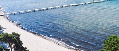 A beach with a long pier extending into the water, surrounded by sand and sparse vegetation.