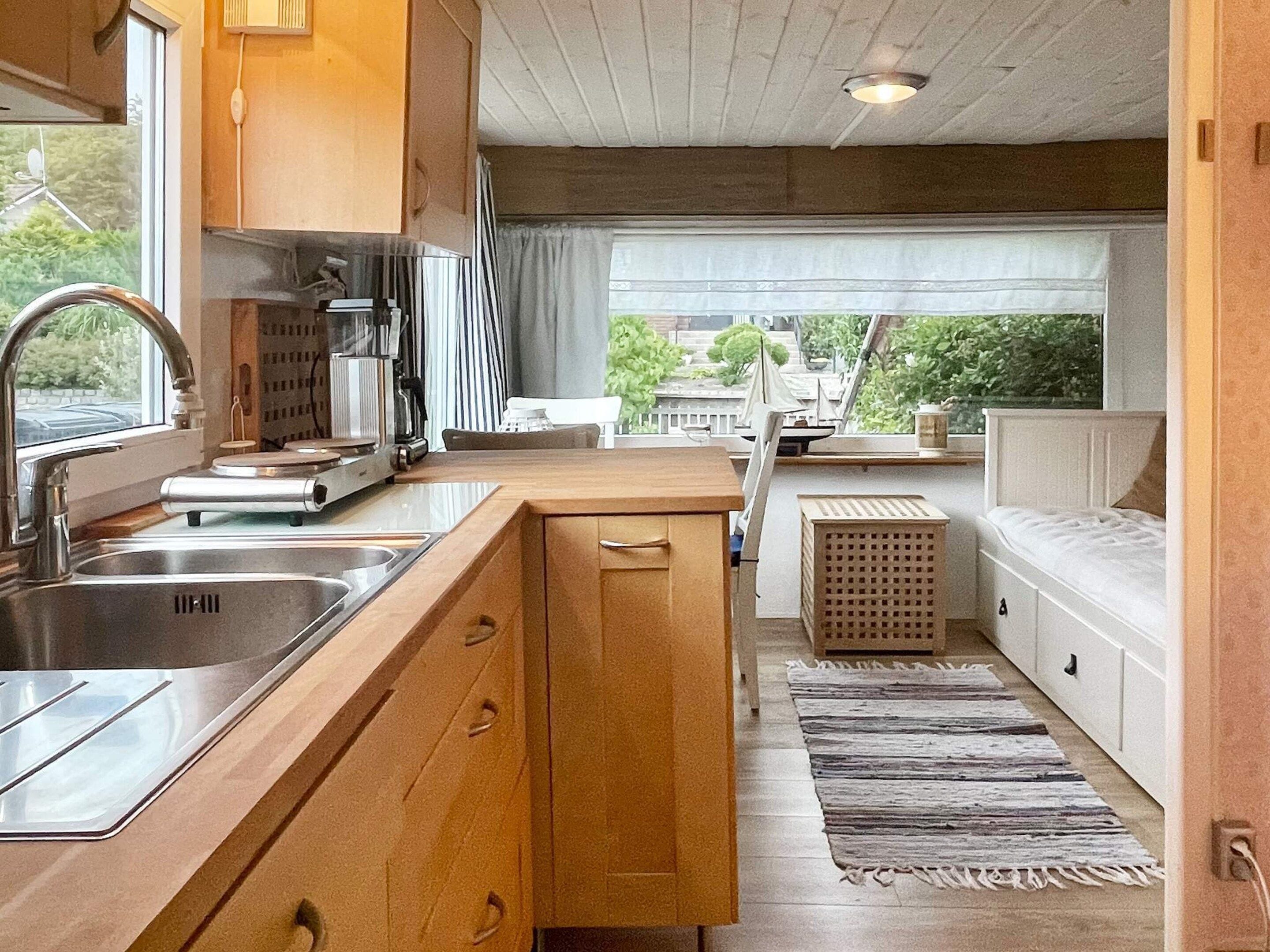 A compact kitchen with a sink, stove, and cabinets, leading to a small dining area with a bench and a view of greenery outside.