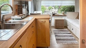 Een kleine keuken met een spoelbak, fornuis en kastjes, die uitkomt op een eethoekje met een bank en uitzicht op groen.