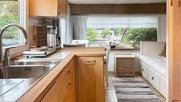 A compact kitchen with a sink, stove, and cabinets, leading to a small dining area with a bench and a view of greenery outside.