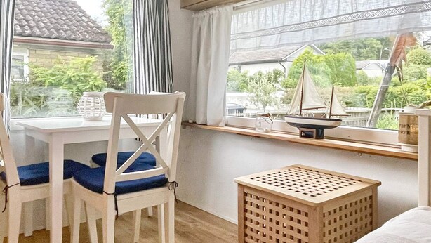 Een kamer met een houten vloer, een witte stoel met een blauw kussen, een klein houten tafeltje, een raam met uitzicht op groen en een zeilboot, en een wit bed.