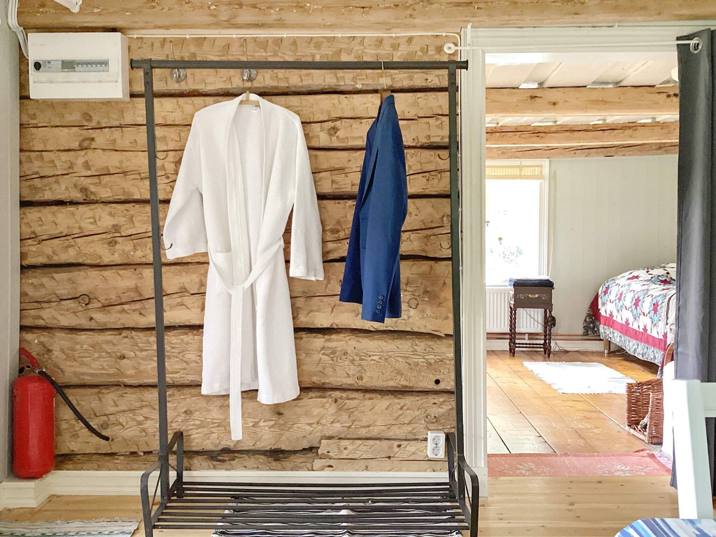 A wooden cabin interior with a metal rack holding two robes, a red fire extinguisher, and a bed with a patterned quilt.