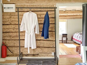 A wooden cabin interior with a metal rack holding two robes, a red fire extinguisher, and a bed with a patterned quilt.