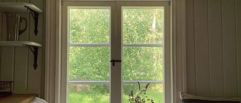 A room with a double door leading to a green outdoor area, a bedside table with a lamp, and a window with a potted plant.