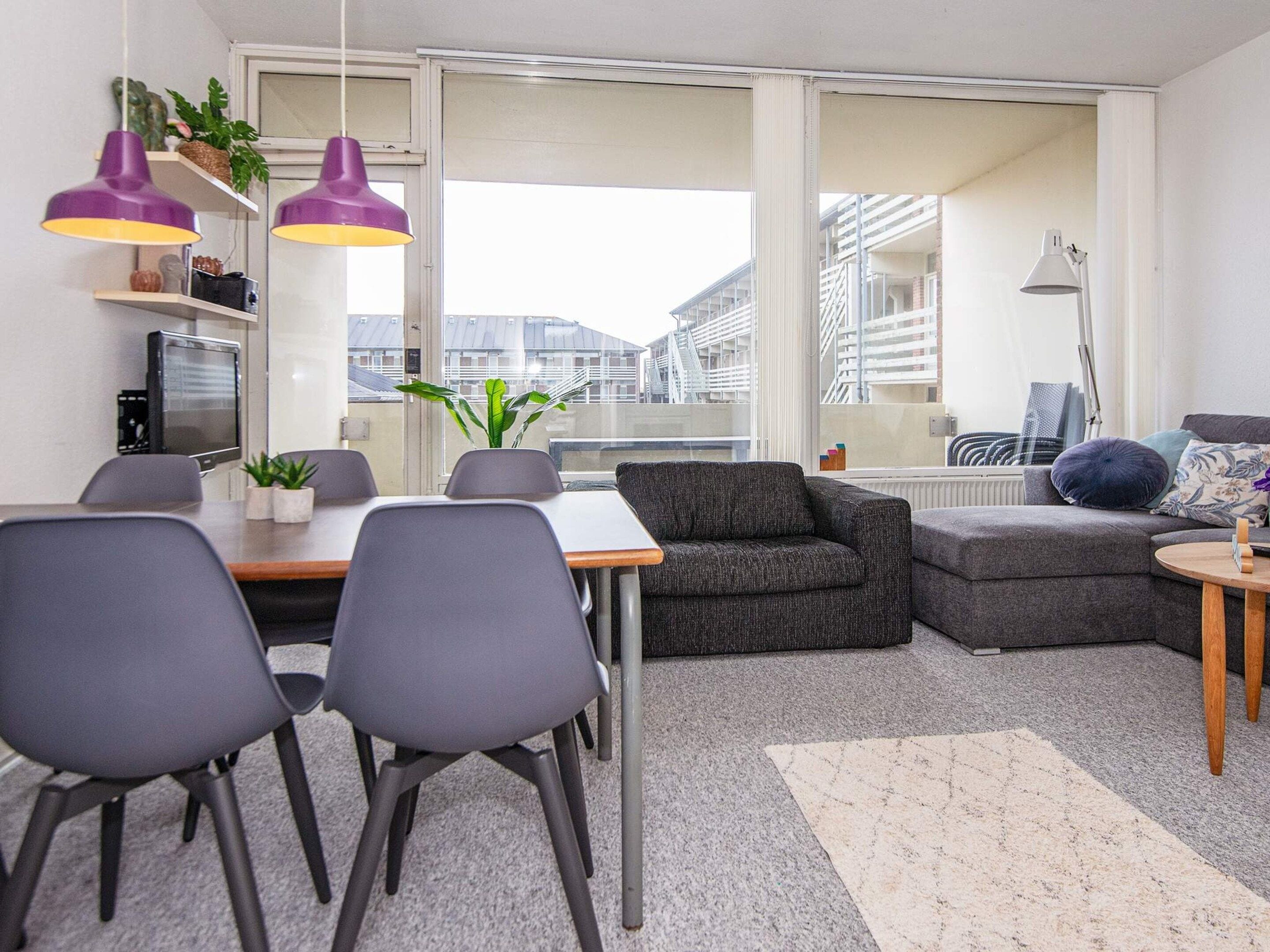 Un salon moderne comprenant un coin repas, des canapés gris, une table basse et une vue sur les immeubles à travers de grandes fenêtres.