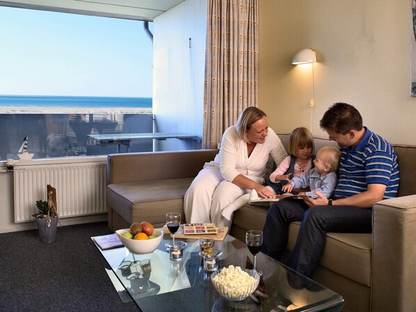 A family sitting on a couch with a view of the ocean, a glass table with snacks and drinks, and a window with curtains. - 6 Person Holiday Park Home in Fano (Fano)