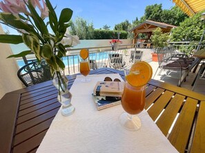 Une table avec des boissons, un livre et un vase de fleurs, le tout placé sur un balcon donnant sur une piscine.