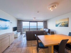 Un salon moderne avec une table à manger en bois, des chaises noires, un canapé en cuir et une grande fenêtre donnant sur la mer.