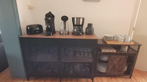 Una kitchenette con cafetera, tostadora y jarra de vidrio sobre la encimera. Hay platos y tazas en un estante debajo.