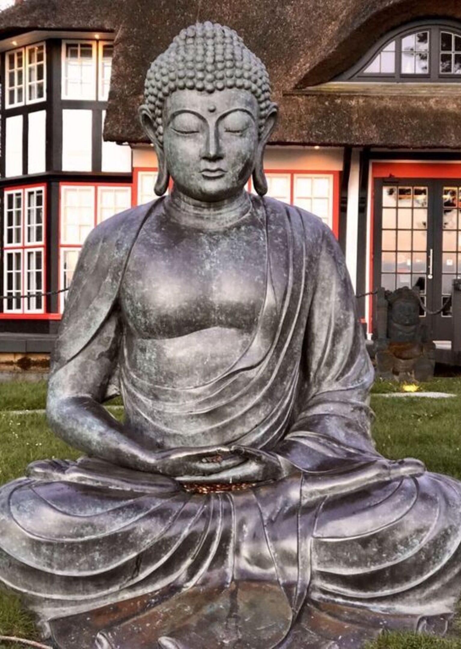 Une grande statue d’extérieur représentant une figure assise dans une pose méditative, avec en arrière-plan un bâtiment traditionnel doté d’un toit de chaume et de détails en rouge.