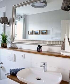 Un baño con un espejo grande, un lavabo blanco, un perchero para toallas, una pequeña planta en maceta y cuadros en la pared.