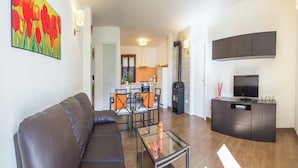 Un salon moderne avec un canapé, une table basse en verre et un téléviseur. Au fond, on aperçoit une cuisine équipée d’une cuisinière et d’un coin repas.