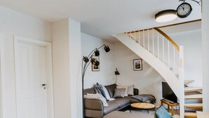 A modern living room with a sofa, a round coffee table, and a chair. There is a staircase with a white railing and a clock on the wall. - Princess Street No8 Comfortable Holiday Residence (Friedrichstadt)