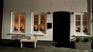 A white building with three windows, each with a candle inside. There is a bench in front of the building and a planter with plants to the right. - Princess Street No8 Comfortable Holiday Residence (Friedrichstadt)