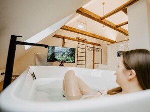 A person relaxing in a bathtub with a television displaying a forest scene, wooden beams, and a brick wall in the background. - Princess Street No8 Comfortable Holiday Residence (Friedrichstadt)