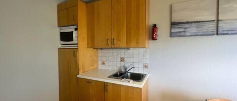 Eine kleine Küche mit Holzschränken, einer weißen Arbeitsplatte, einem Edelstahl-Spülbecken und einer Mikrowelle.