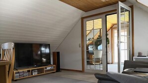 A modern living room with a flat-screen TV, wooden entertainment unit, and a sliding glass door leading to a balcony.