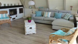 Ein gemütliches Wohnzimmer mit Kamin, einem grauen Sofa, Korbstühlen und einem kleinen weißen Tisch mit einem Topf einer Pflanze.