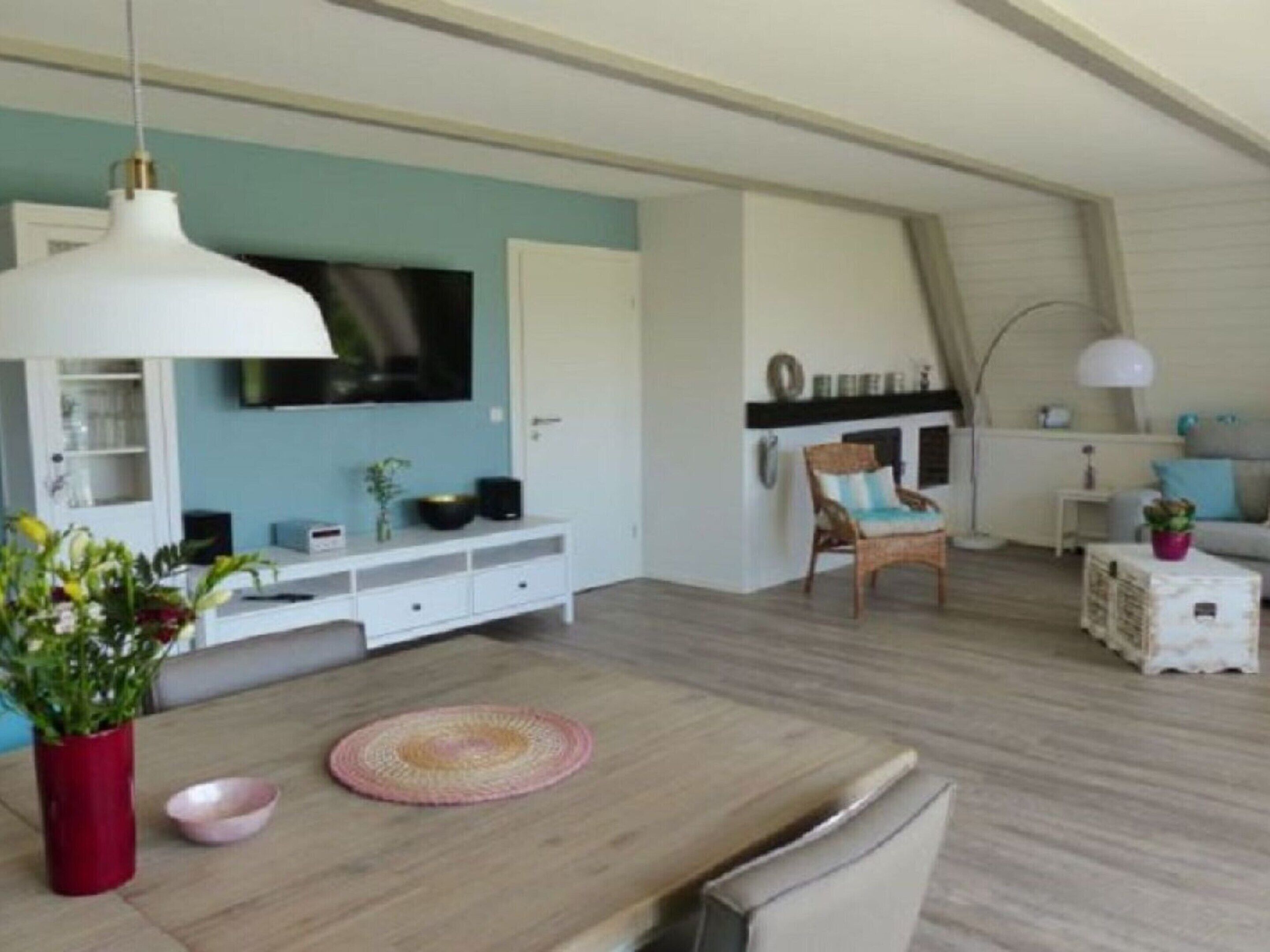 Una sala moderna con una mesa de comedor de madera, un televisor montado en la pared y una zona de estar acogedora con una silla y una mesita auxiliar.
