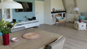 Ein modernes Wohnzimmer mit einem Holzesstisch, einem an der Wand montierten Fernseher und einer gemütlichen Sitzecke mit einem Sessel und einem kleinen Beistelltisch.