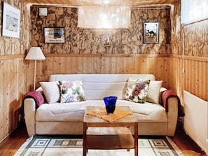 Una habitación con un panel de madera, un sofá blanco con cojines florales, un jarrón azul sobre una mesa de madera y una alfombra estampada.