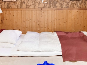 Una cama con ropa blanca y una manta marrón, frente a una pared de madera con textura.